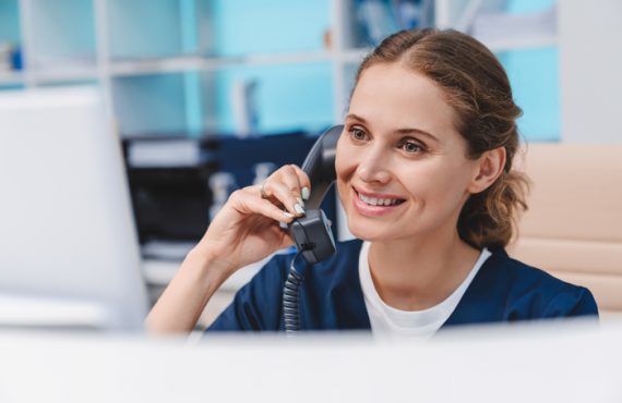 Young female nurse working at reception desk while answering phone calls and scheduling appointments
