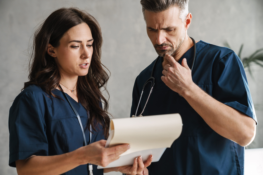 Two serious female and male nurse triage in medical uniform looking at clipboard while standing indoors